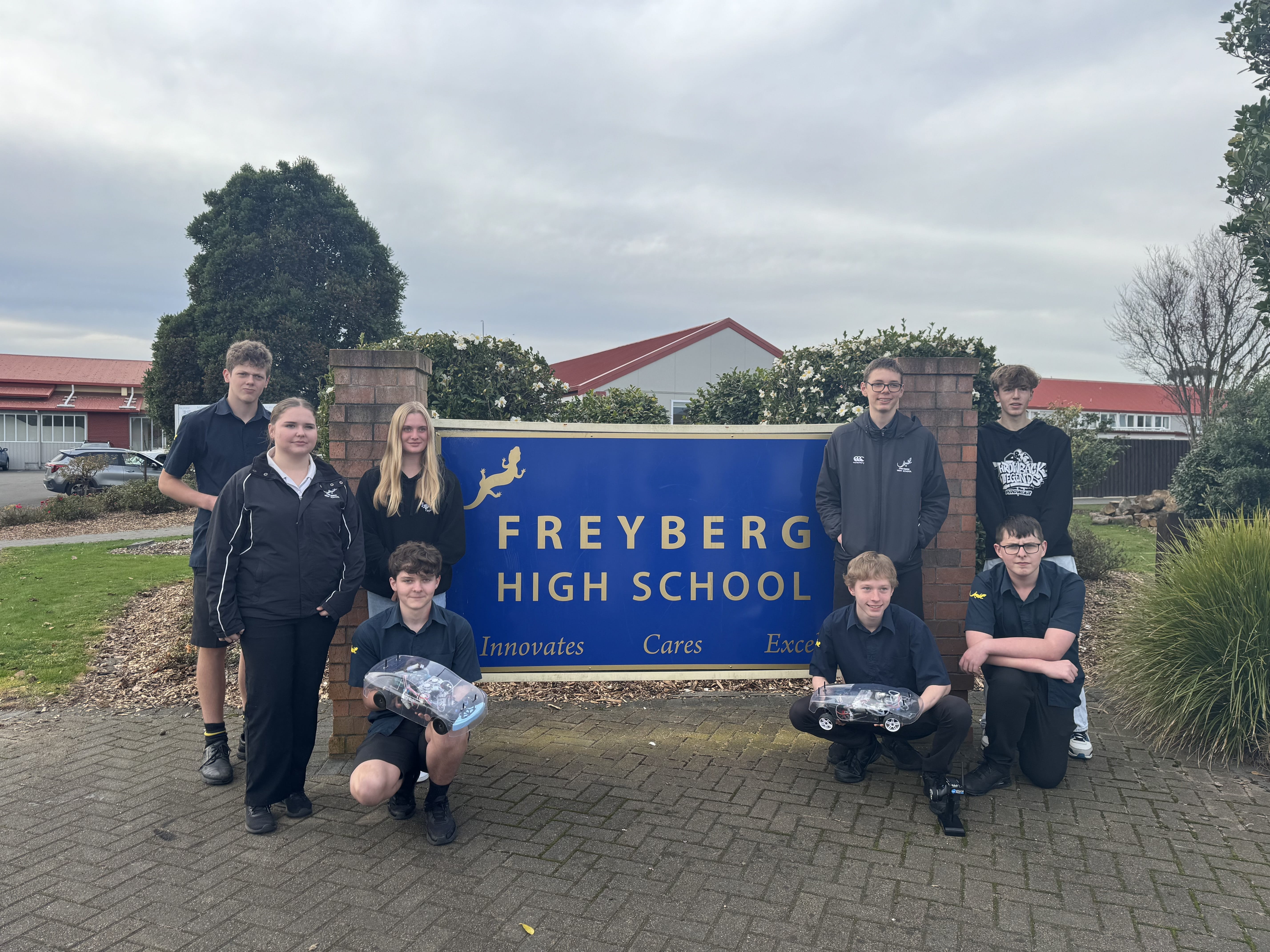 Eight young people from Freyberg High School gathered around their school's sign. Two boys in the front are holding hydrogen remote control cars.