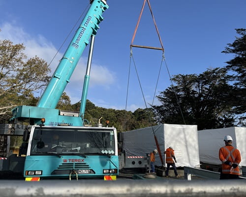 A container sized object wrapped in white plastic is lowered to the ground by a crane 