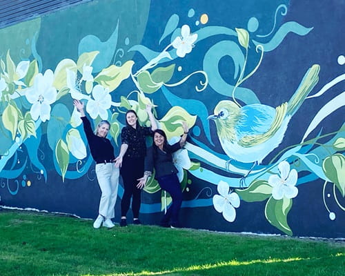 Three women with their hands in the air in front of a mural on a outside wall, depicting kiwifruit vines and flowers, and a wax-eye bird