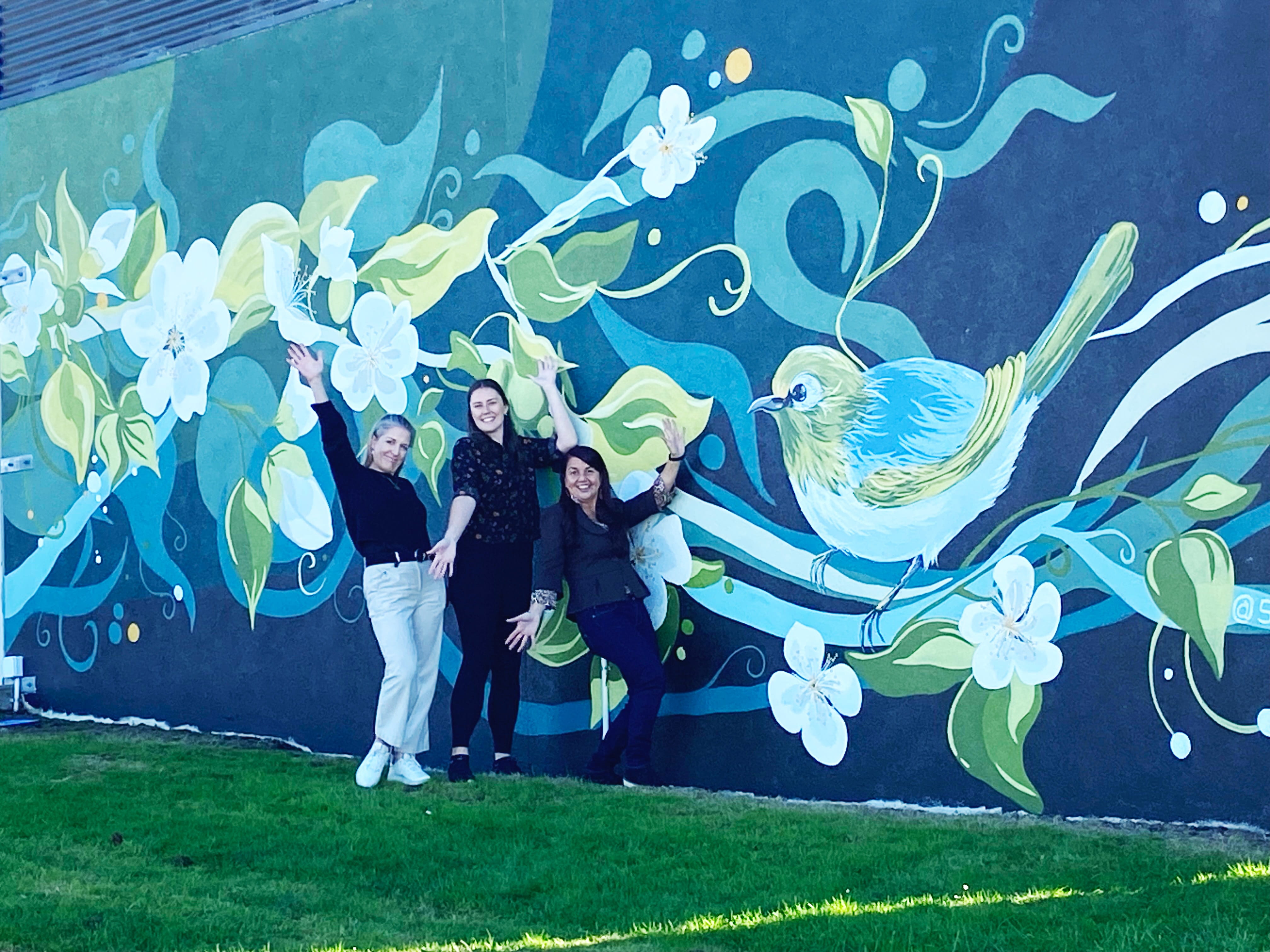 Three women with their hands in the air in front of a mural on wall depicting kiwifruit vine, flowers and a wax-eye bird.